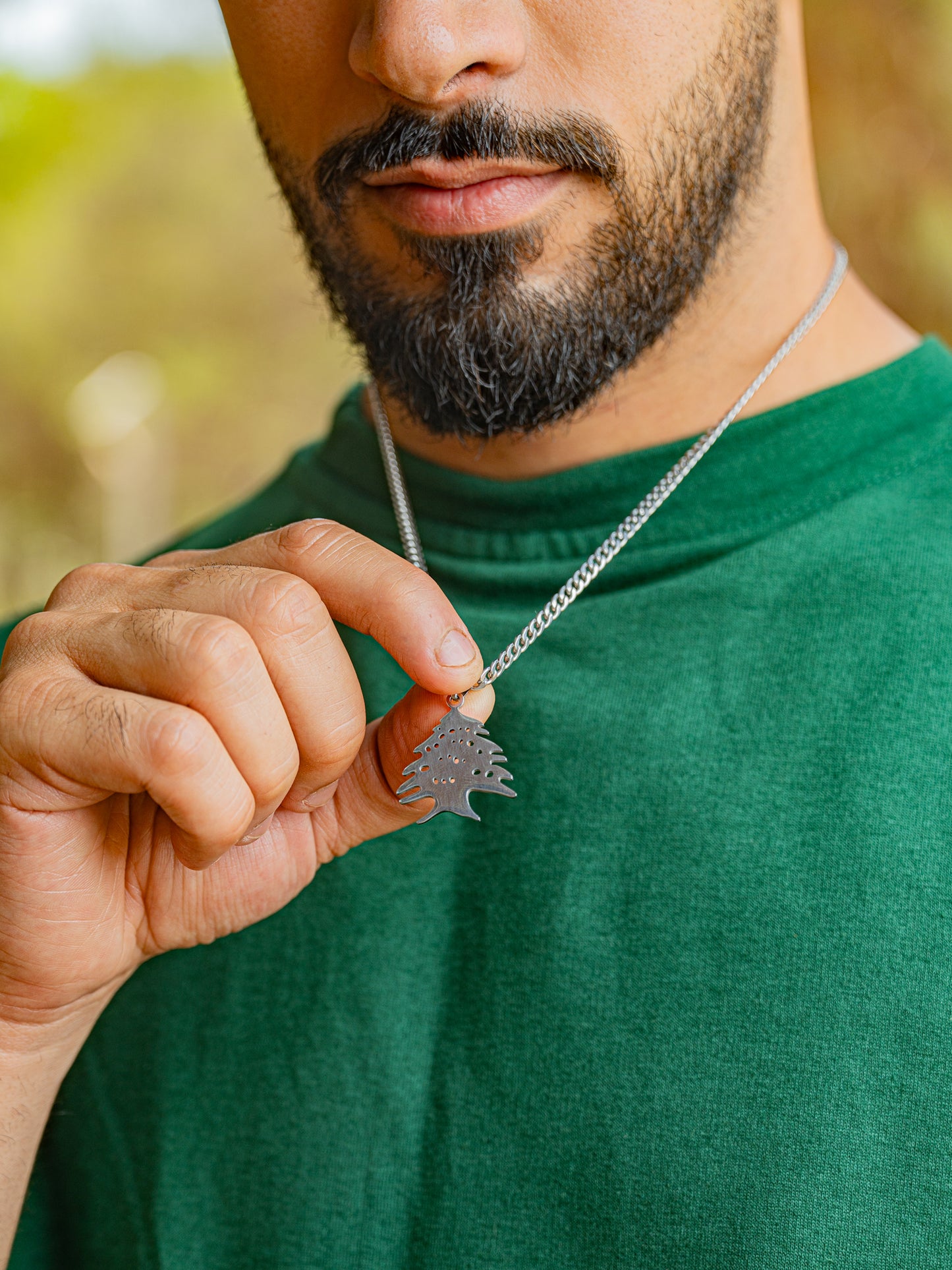 Man holding a silver necklace with a lebanese cedar tree icon pendant against a blurred natural background