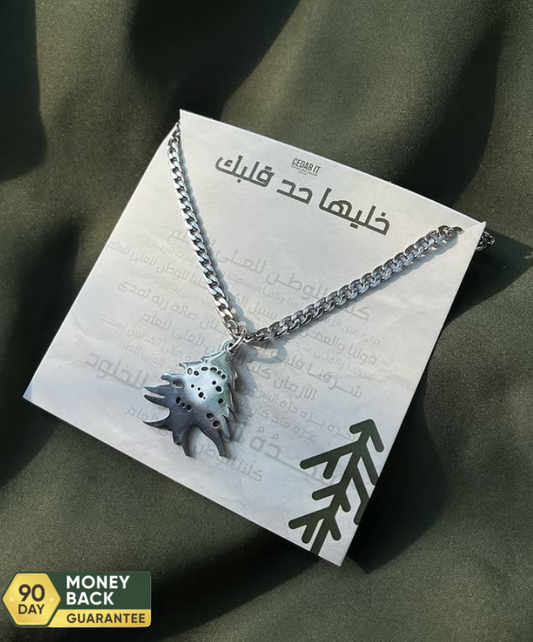 A stainless steel necklace with a pendant in the shape of the Cedar of Lebanon, displayed on a card with Arabic script.